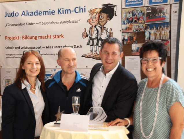 Vier Personen posieren l&auml;chelnd an einem Stehtisch vor einem Banner der Judo Akademie Kim-Chi.