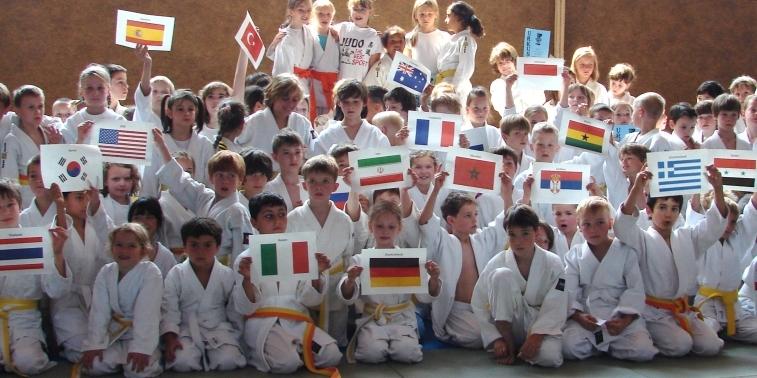 Gruppierung von Kindern in Judogis, die unterschiedliche L&auml;nderflaggen hochhalten, auf einer Judo-Matte.