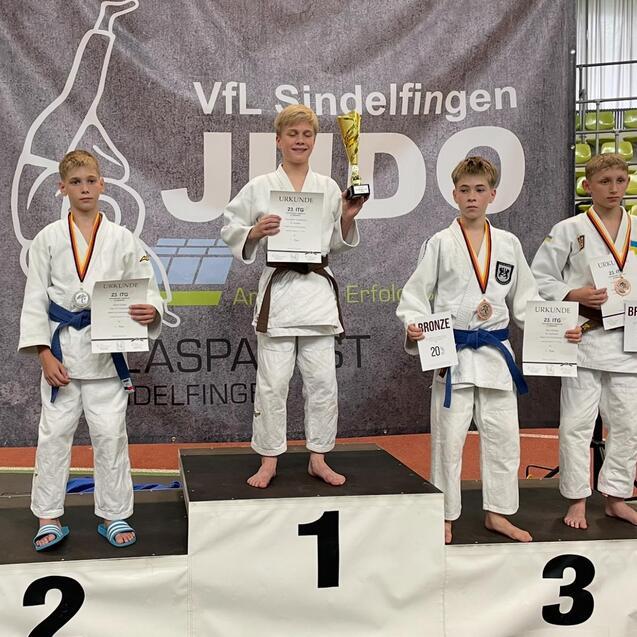 Vier Judokas stehen auf dem Siegertreppchen, w&auml;hrend der zentrale K&auml;mpfer den Pokal und eine Urkunde h&auml;lt.