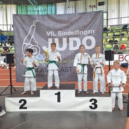 Vier Kinder in Judoanz&uuml;gen stehen auf dem Siegertreppchen, halten Troph&auml;en und Urkunden, mit einem Judo-Logo im Hintergrund.