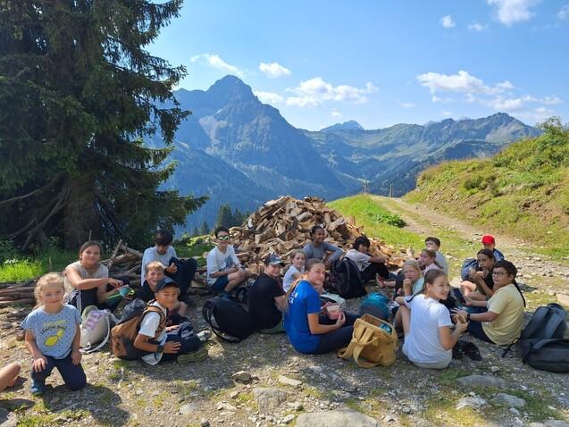 Gruppe von Kindern und Jugendlichen sitzt auf einem Weg in den Bergen, im Hintergrund sind B&auml;ume und Berge zu sehen.