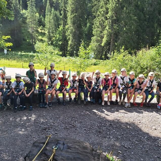 Gruppe von Kindern und Betreuern in Kletterausr&uuml;stung und Helmen, sitzend in der Natur.