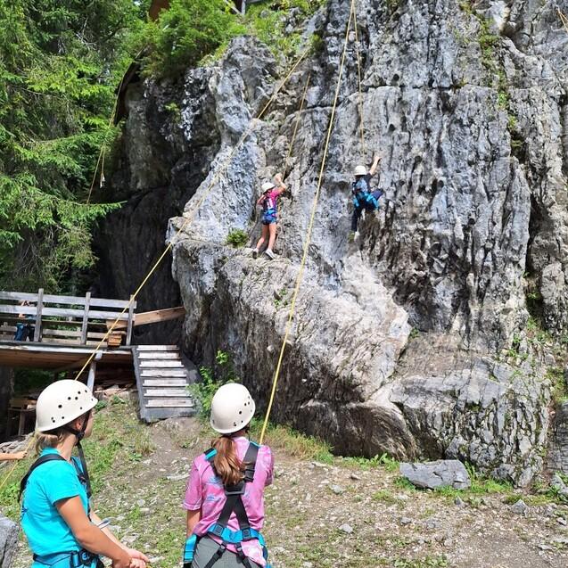 Drei Kinder in Kletterausr&uuml;stung sichern zwei Kletterer an einer steilen Felswand im Wald.