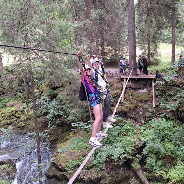 Zwei Personen stehen auf einer Seilbr&uuml;cke zwischen B&auml;umen &uuml;ber einem Bach, im Hintergrund weitere Personen und Natur.