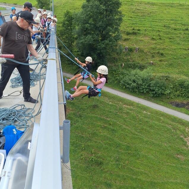 Gruppen von Personen sichern Kinder auf einem Hochseil mit Kletterausr&uuml;stung &uuml;ber einer Wiese in Berglandschaft.