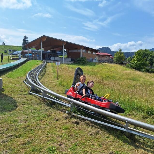 Zwei Kinder sitzen in einer roten Sommerrodelbahn, w&auml;hrend die Strecke durch eine gr&uuml;ne Wiese verl&auml;uft.