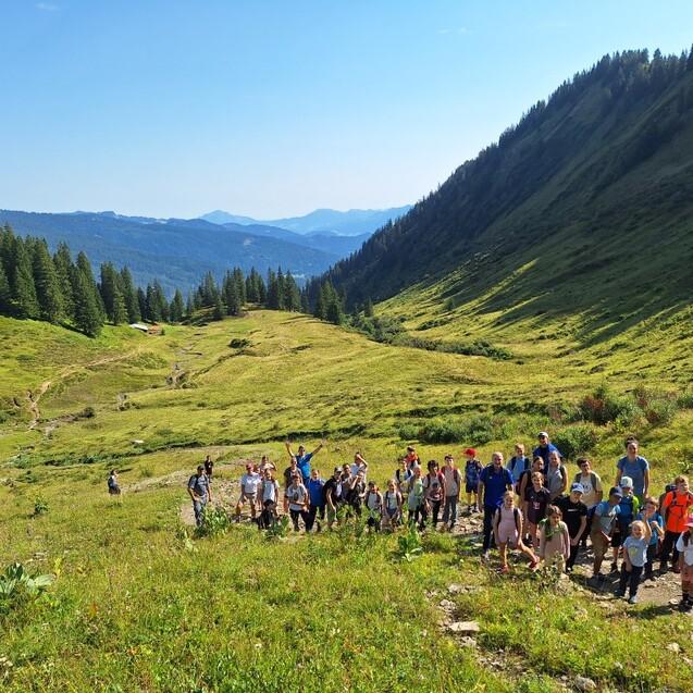 Gruppe von Wanderern auf einem gr&uuml;nen Hang mit B&auml;umen im Hintergrund und Bergen unter blauem Himmel.