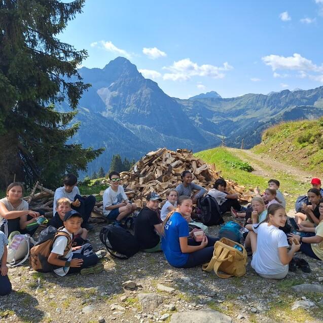 Gruppe von Kindern und Jugendlichen sitzt auf einem Weg in den Bergen, im Hintergrund sind B&auml;ume und Berge zu sehen.