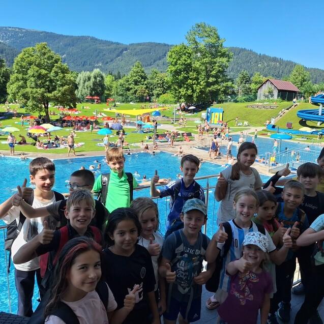 Gruppe von Kindern mit Rucks&auml;cken am Rand eines Schwimmbads, im Hintergrund sind Rutschen und sonnige Wiesen.