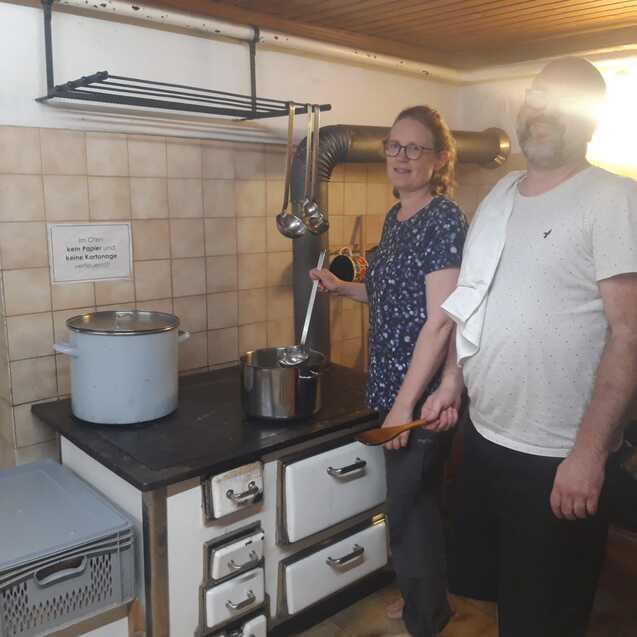 Zwei Personen in einer K&uuml;che stehen an einem Herd mit einem Topf und einer Kelle in der Hand.