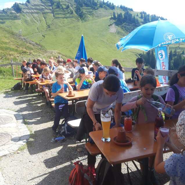 Gruppe von Kindern und Jugendlichen sitzt an Tischen im Freien, mit Blick auf eine gr&uuml;ne Berglandschaft.