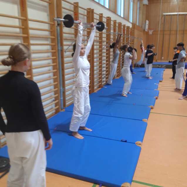 Gruppentraining in einer Sporthalle: Jugendliche heben Gewichte auf blauen Matten, w&auml;hrend Trainerin beobachtet.