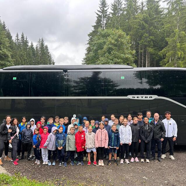 Gruppe von Kindern und Betreuern posiert vor einem gro&szlig;en schwarzen Reisebus in einem Waldgebiet.