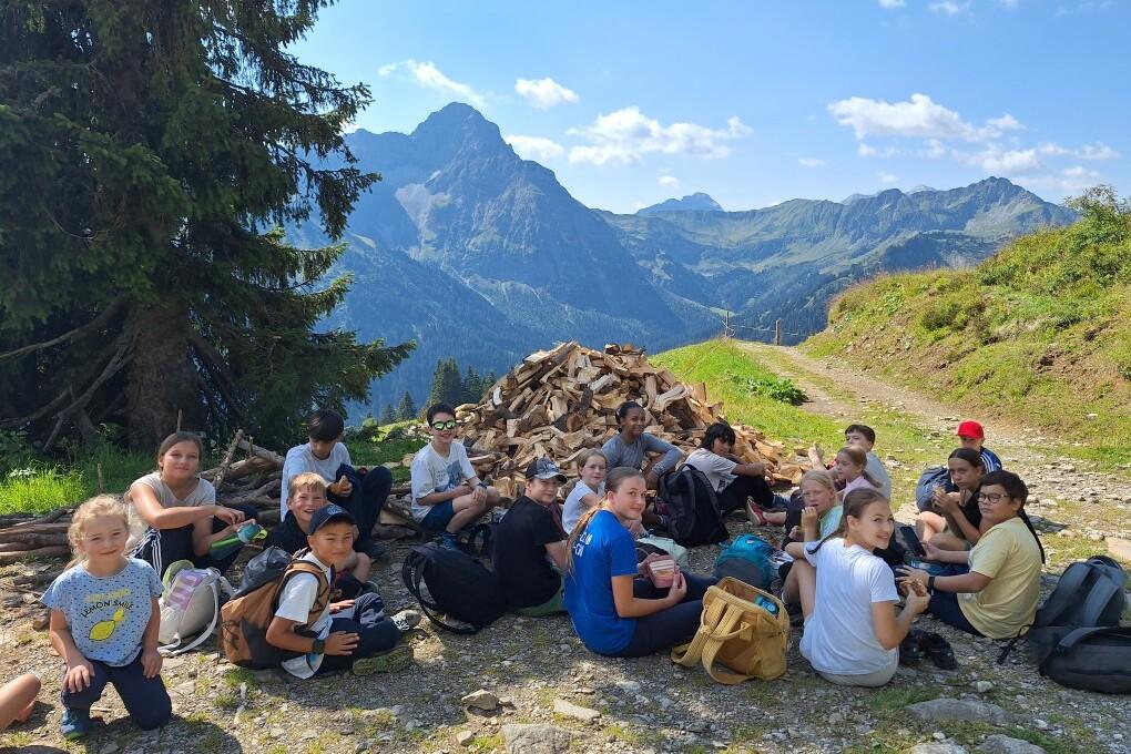 Gruppe von Kindern und Jugendlichen sitzt auf einem Weg in den Bergen, im Hintergrund sind B&auml;ume und Berge zu sehen.