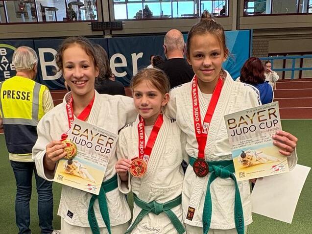 Drei junge Judoka in wei&szlig;en Anz&uuml;gen mit Medaillen und Urkunden beim Bayer Judo Cup auf einer Sportfl&auml;che.