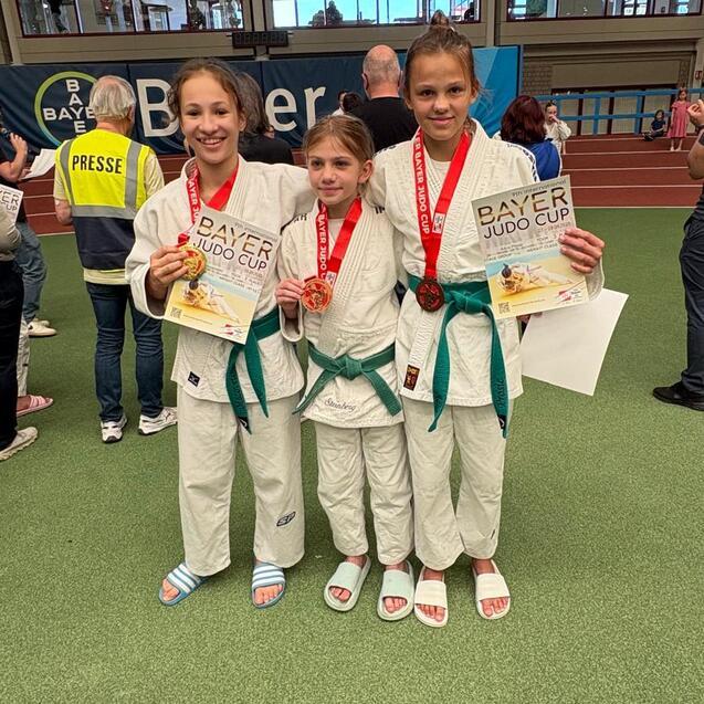 Drei junge Judoka in wei&szlig;en Anz&uuml;gen mit Medaillen und Urkunden beim Bayer Judo Cup auf einer Sportfl&auml;che.