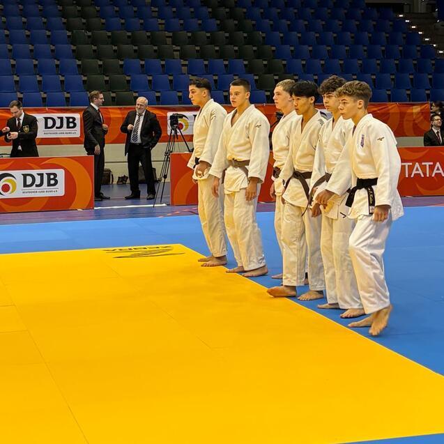 Sechs Judokas in wei&szlig;en und blauen Judogis stehen auf einer Mattenfl&auml;che, bereit f&uuml;r den Wettkampf.