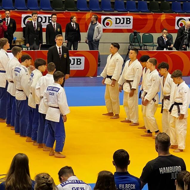 Zwei Gruppen von Judokas in wei&szlig;en und blauen Anz&uuml;gen stehen auf einer Judomatte, beobachtet von Zuschauern.