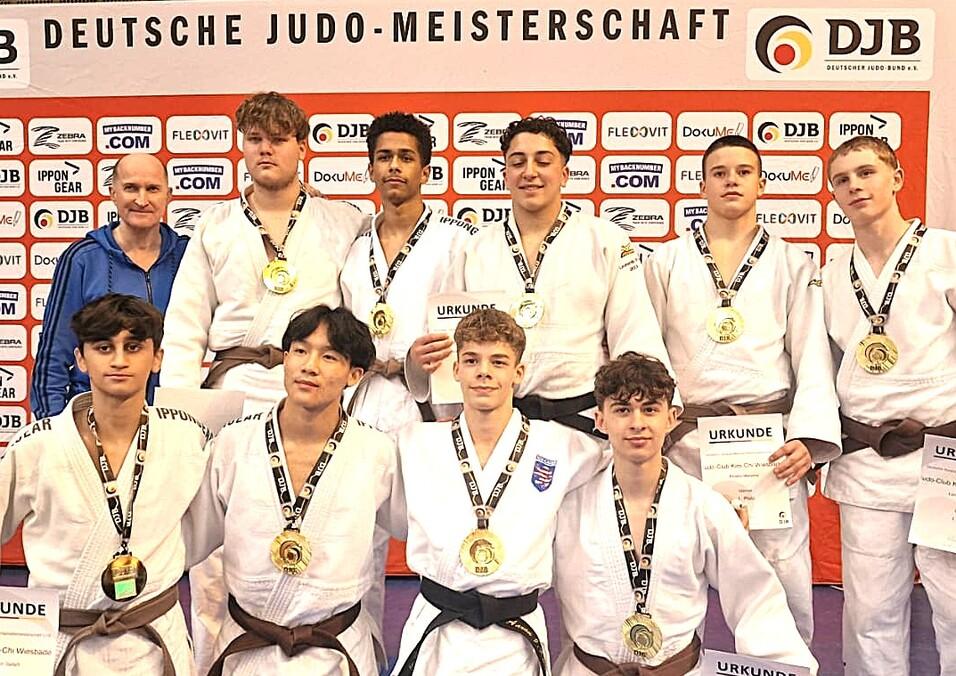 Gruppe von Judoka in weißen Gi und braunen Gürteln, posierend mit Medaillen und Urkunden vor einem Banner der Deutschen Judo-Meisterschaft.