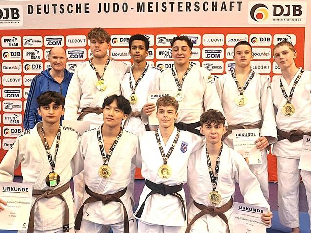 Gruppe von Judoka in wei&szlig;en Gi und braunen G&uuml;rteln, posierend mit Medaillen und Urkunden vor einem Banner der Deutschen Judo-Meisterschaft.