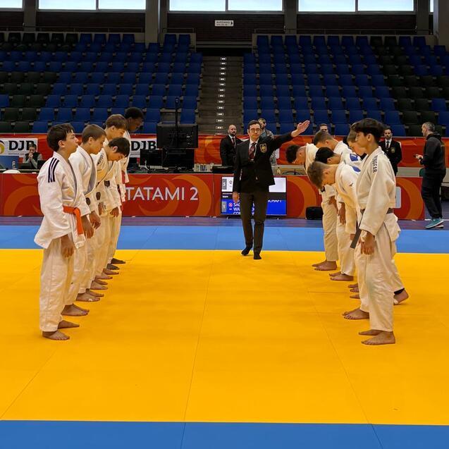 Zwei Gruppen junger Judokas stehen sich auf einer blauen Matte gegen&uuml;ber, w&auml;hrend ein Schiedsrichter im Hintergrund Anweisungen gibt.