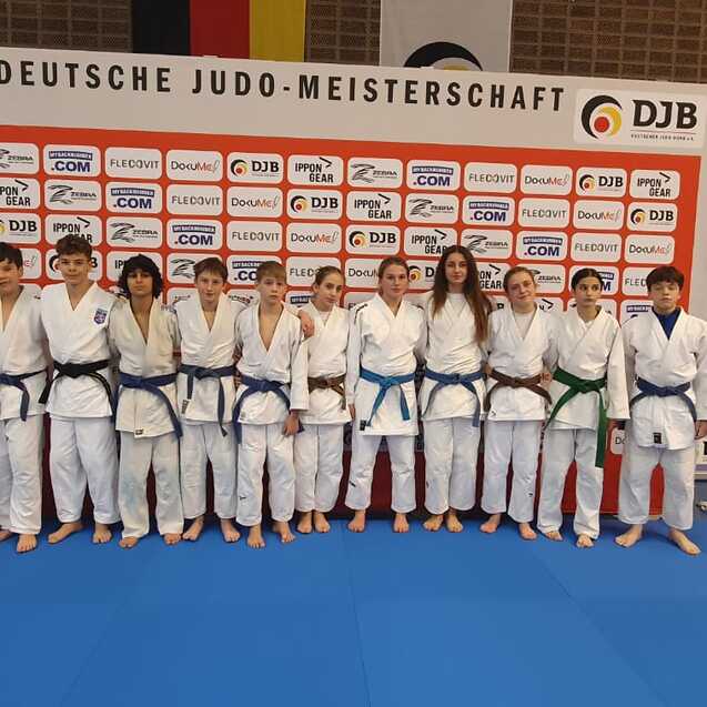 Gruppe von Judokas in wei&szlig;en Anz&uuml;gen und blauen G&uuml;rteln steht vor einer Mauer mit Sponsorenlogos bei der Deutschen Judo-Meisterschaft.