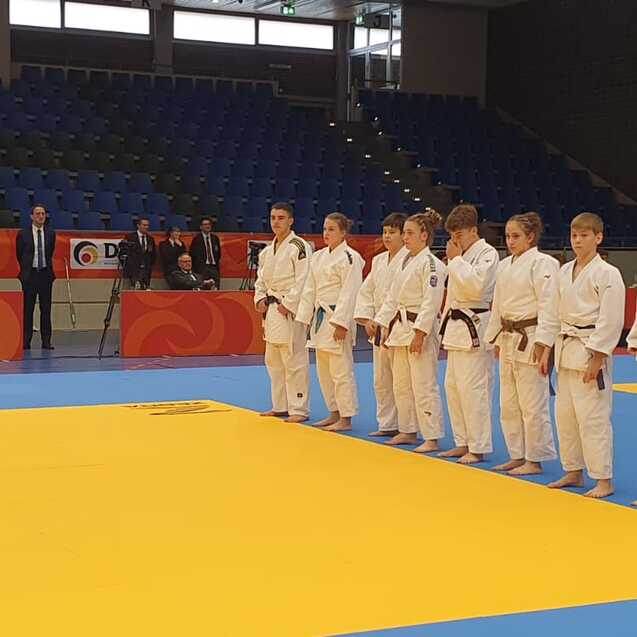 Acht Judoka in wei&szlig;en Kimonos stehen in Reihe auf einer Judomatte, w&auml;hrend ein Schiedsrichter sie beobachtet.