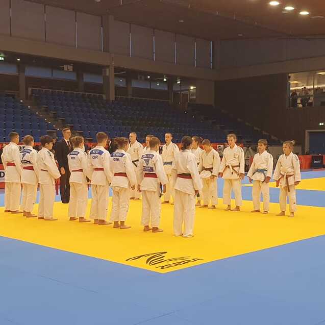 Gruppierung von Judokas in wei&szlig;en Anz&uuml;gen, die auf einer gelben Judomatte in einer Sporthalle stehen.
