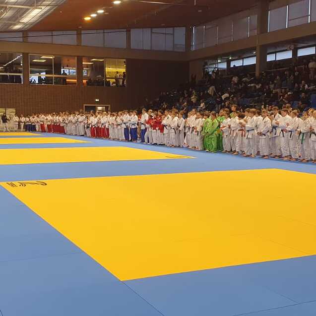 Vielzahl von Karateka in Uniform versammelt auf blauen Matten w&auml;hrend einer Wettkampfveranstaltung in einer Sporthalle.