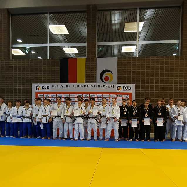 Gruppenfoto von Judokas in Judogi mit Medaillen und Urkunden auf einer Wettkampfmatte, im Hintergrund Flaggen und Banner.