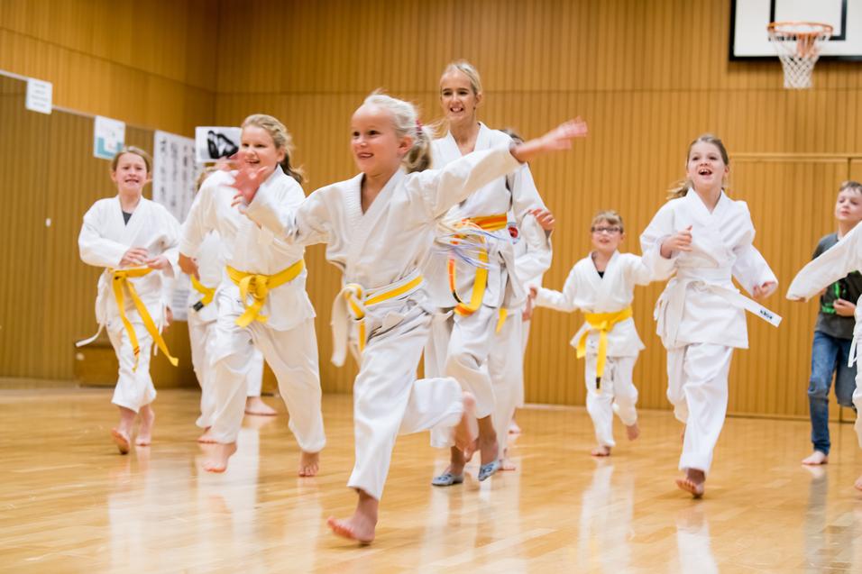Gruppe von Kindern in wei&szlig;en Karateanz&uuml;gen mit gelben G&uuml;rteln, die fr&ouml;hlich in einer Sporthalle laufen.