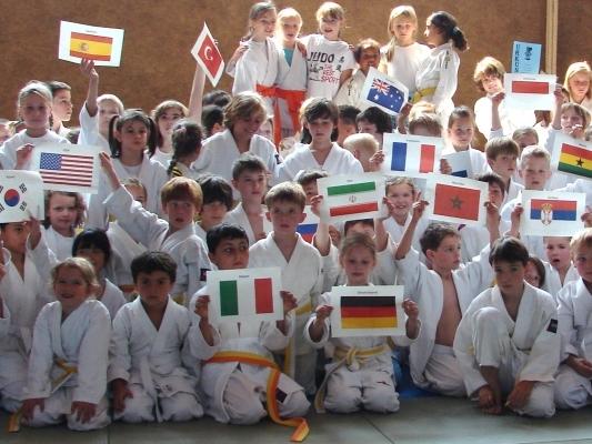 Gruppierung von Kindern in Judogis, die unterschiedliche L&auml;nderflaggen hochhalten, auf einer Judo-Matte.