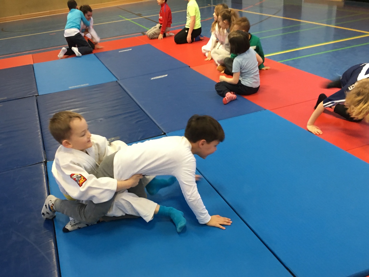 Zwei Kinder im Judoanzug &uuml;ben verschiedene Techniken auf blauen und roten Matten in einer Sporthalle.