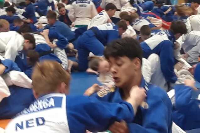 Gruppentraining von Judokas in blauen und wei&szlig;en Anz&uuml;gen auf Matten in einer Sporthalle. Zuschauer im Hintergrund.