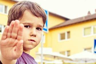 Junge h&auml;lt Hand hoch zur Sicherheit am Schulweg, im Hintergrund sind Geb&auml;ude und Verkehrsschilder sichtbar.