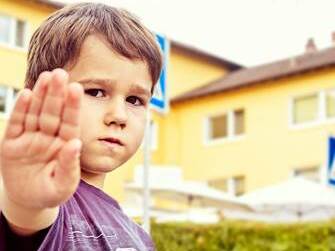 Junge h&auml;lt Hand hoch zur Sicherheit am Schulweg, im Hintergrund sind Geb&auml;ude und Verkehrsschilder sichtbar.