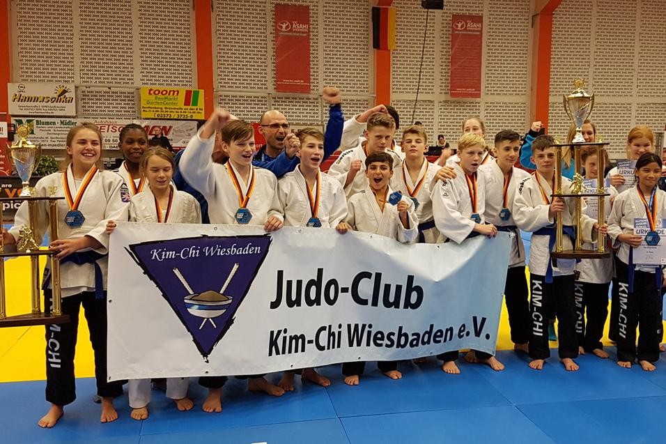 Gruppe junger Judoka des Kim-Chi Wiesbaden e.V. posiert stolz mit Pokalen und Medaillen auf einer Judo-Wettkampffl&auml;che.