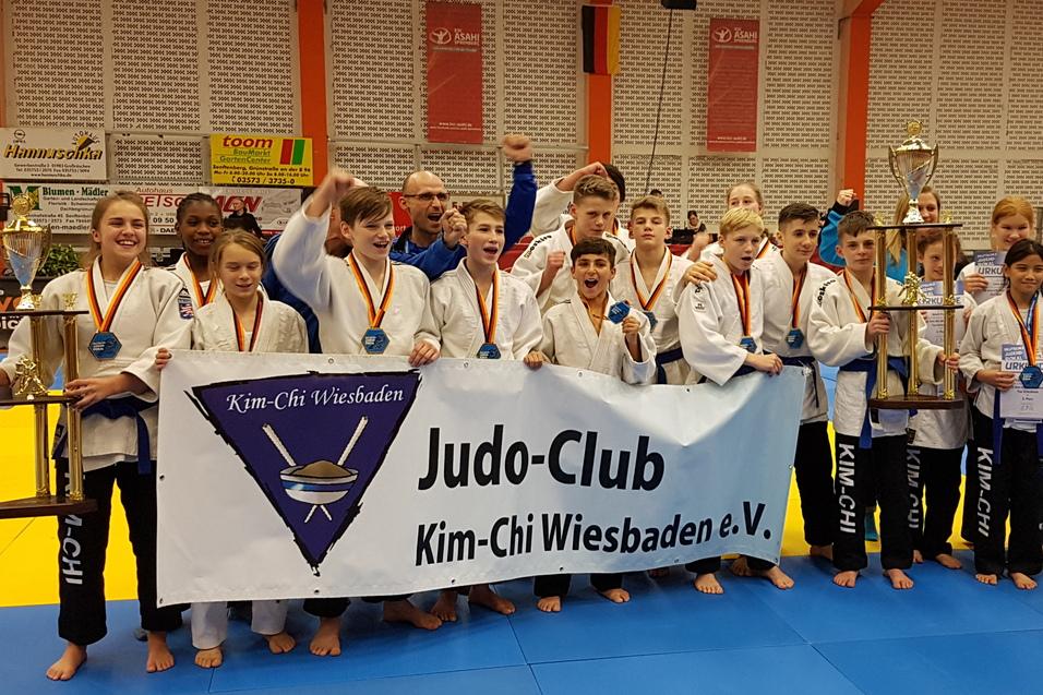 Gruppe junger Judoka des Kim-Chi Wiesbaden e.V. posiert stolz mit Pokalen und Medaillen auf einer Judo-Wettkampffl&auml;che.