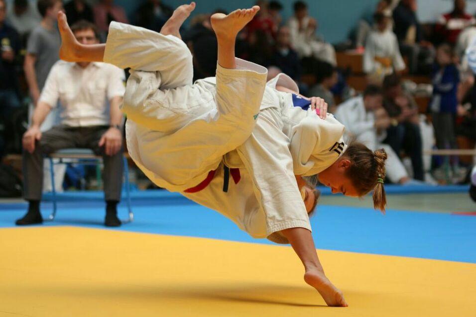 Junge Judoka f&uuml;hrt einen Wurf aus, w&auml;hrend sie auf einer blauen und gelben Judomatte in einem Wettkampf steht.