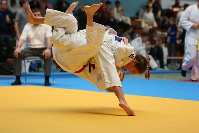 Junge Judoka f&uuml;hrt einen Wurf aus, w&auml;hrend sie auf einer blauen und gelben Judomatte in einem Wettkampf steht.