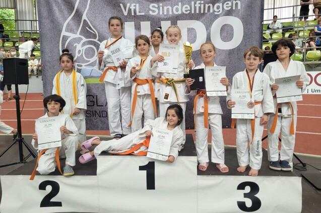 Gruppe von Kindern in bunten Judo-Anz&uuml;gen, die stolz Urkunden und eine Troph&auml;e auf dem Siegerpodest halten.