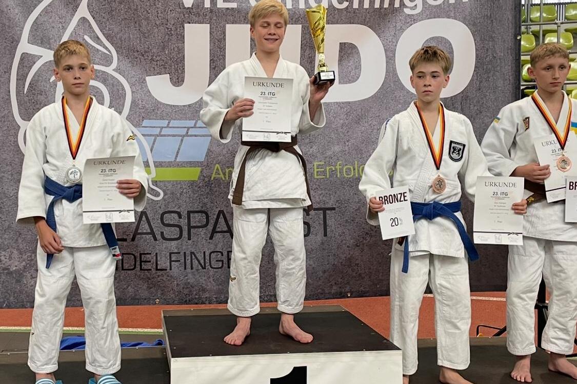 Vier Judoka stehen auf dem Treppchen, der Sieger h&auml;lt einen Pokal und eine Urkunde, die anderen zeigen ihre Medaillen.