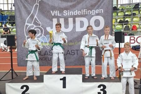 Vier Kinder in Judokleidung stehen auf einem Siegertreppchen, halten Troph&auml;en und Urkunden vor einem Judo-Plakat.