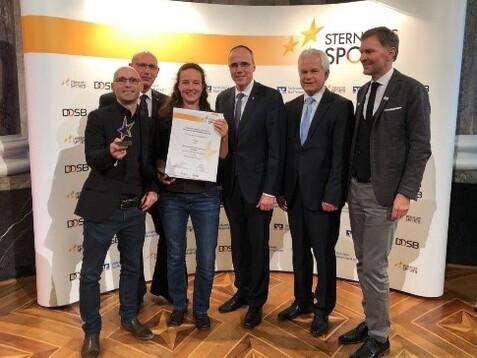 Sechs Personen stehen vor einer Veranstaltungswand und halten eine Auszeichnung und eine Urkunde.