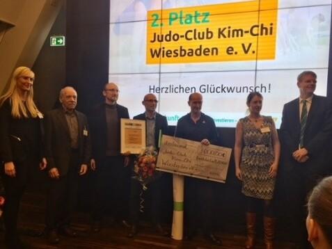 Sieben Personen stehen auf einer Bühne und halten eine Urkunde sowie einen Scheck für den Judo-Club Kim-Chi Wiesbaden.