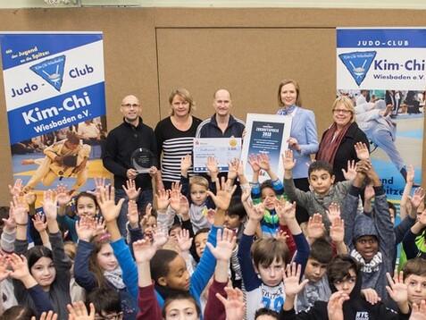 Gruppenfoto mit Kindern und Erwachsenen, die stolz ihre Hände heben, vor Bannern des Judo-Clubs Kim-Chi Wiesbaden.