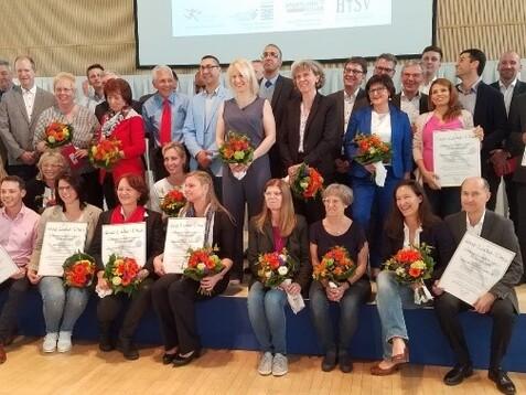 Gruppenfoto von mehreren Personen auf einer Bühne, die Urkunden und Blumenstrauß halten. Veranstaltung mit festlichem Charakter.
