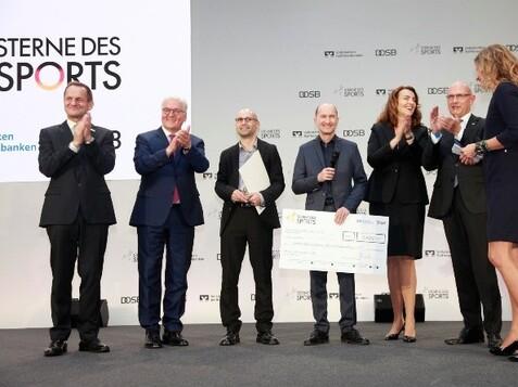 Sechs Personen stehen auf der Bühne, applaudieren und halten ein großes Scheck über eine Auszeichnung in der Hand.