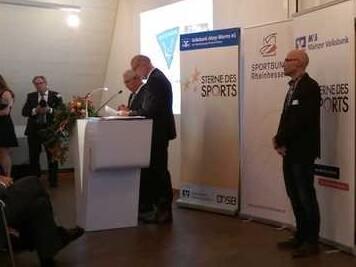 Veranstaltungshalle mit Rednern an einem Podium, Plakate mit „Sterne des Sports“ und Teilnehmenden im Vordergrund.