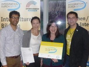 Vier Personen stehen zusammen und halten ein Zertifikat, umgeben von Bannern zur Integration durch Sport.